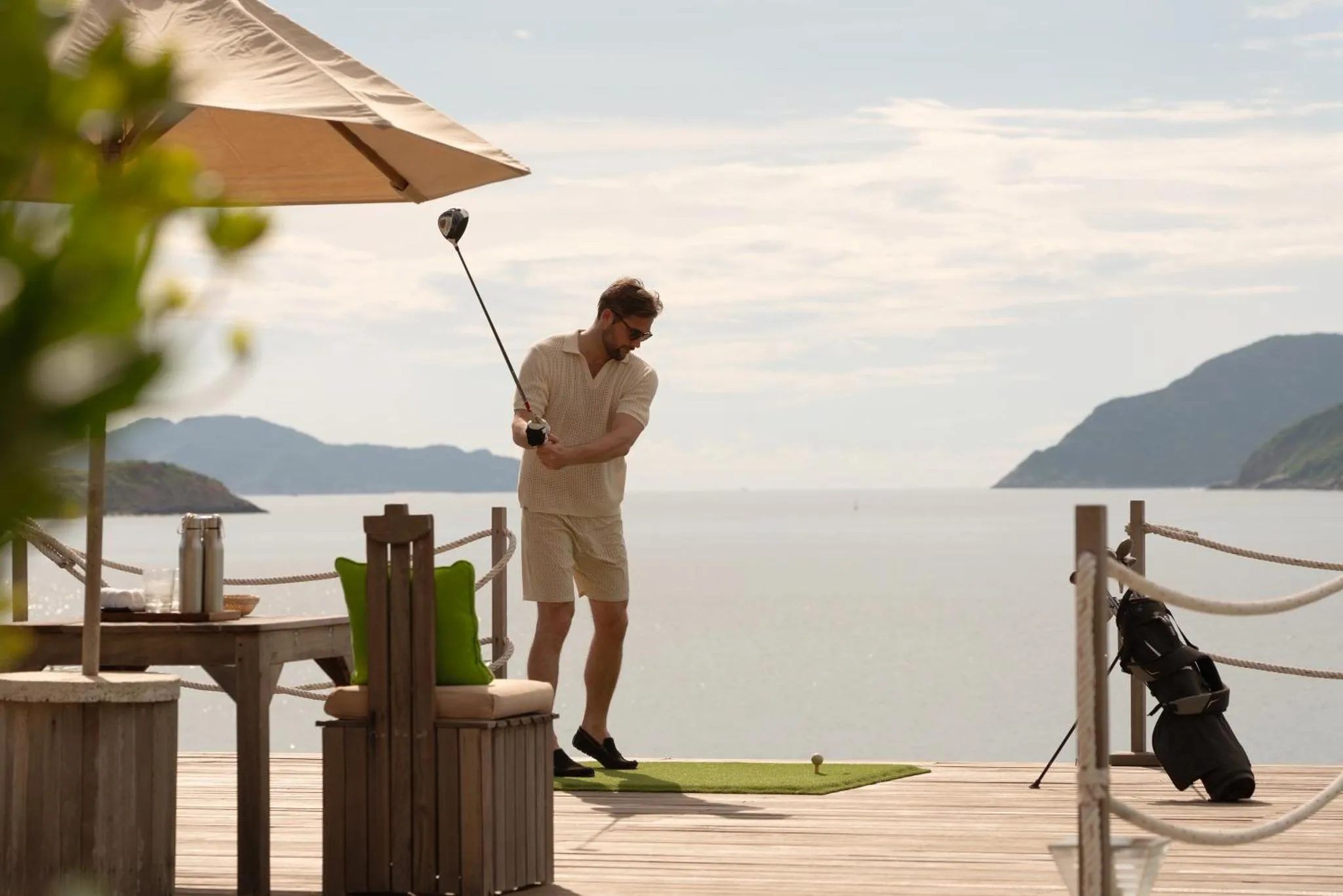 Un homme frappe une balle de golf sur une terrasse en bois avec vue sur la mer.