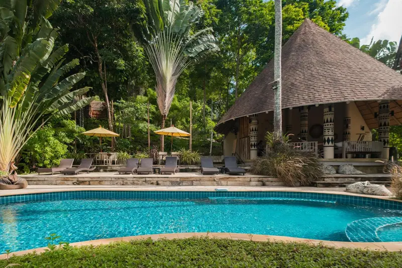 Une piscine turquoise bordée de transats, devant un bâtiment à toit de chaume dans une végétation luxuriante.