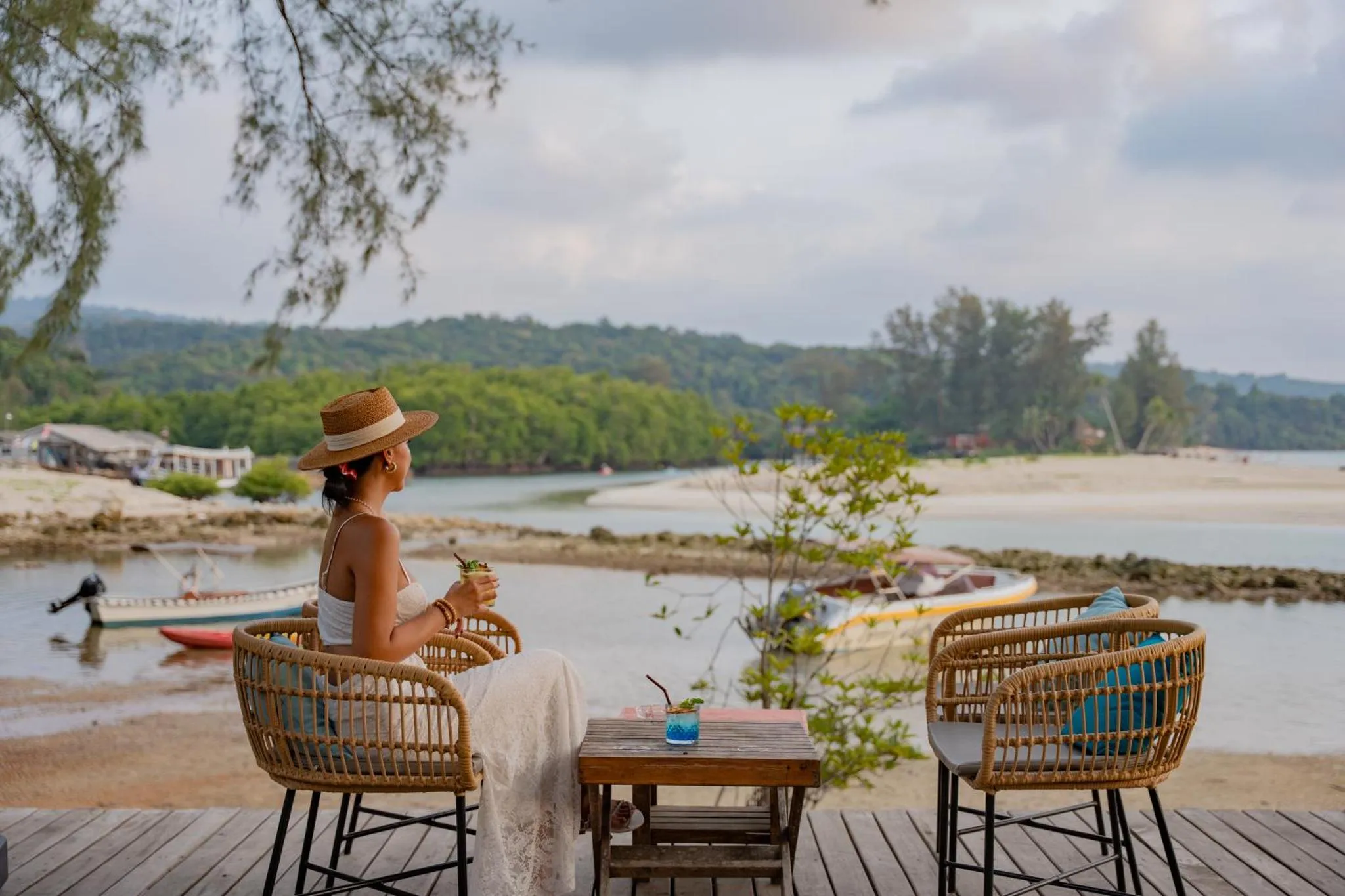Une femme assise à une table en osier avec une boisson, face à la mer.