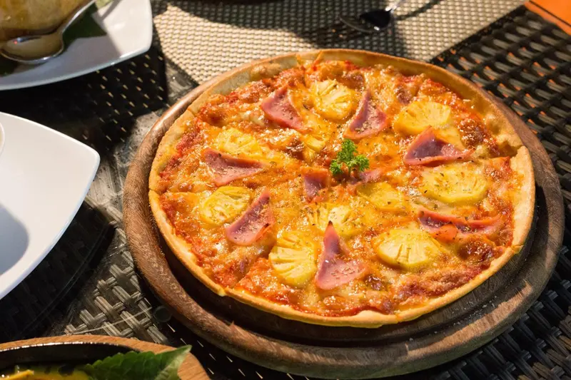 Une pizza garnie d'ananas et de jambon est présentée sur une planche en bois.