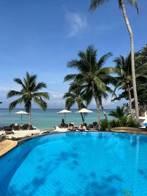 Piscine turquoise bordée de transats sous des palmiers, avec la mer en arrière-plan.