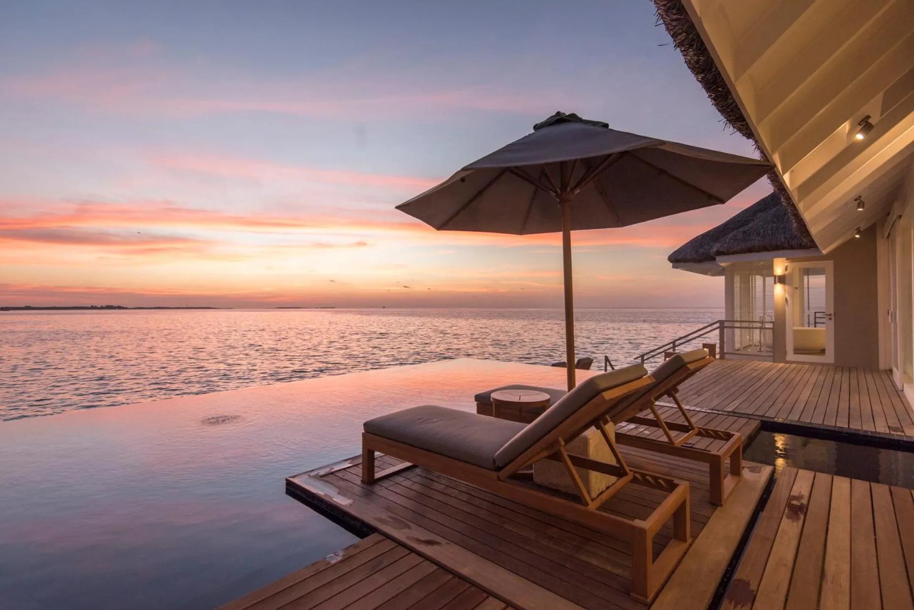 Terrasse en bois avec transats et piscine à débordement face à la mer au coucher du soleil.