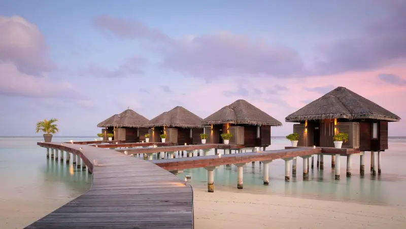 Des bungalows sur pilotis s'alignent le long d'une passerelle en bois au-dessus d'une eau turquoise.