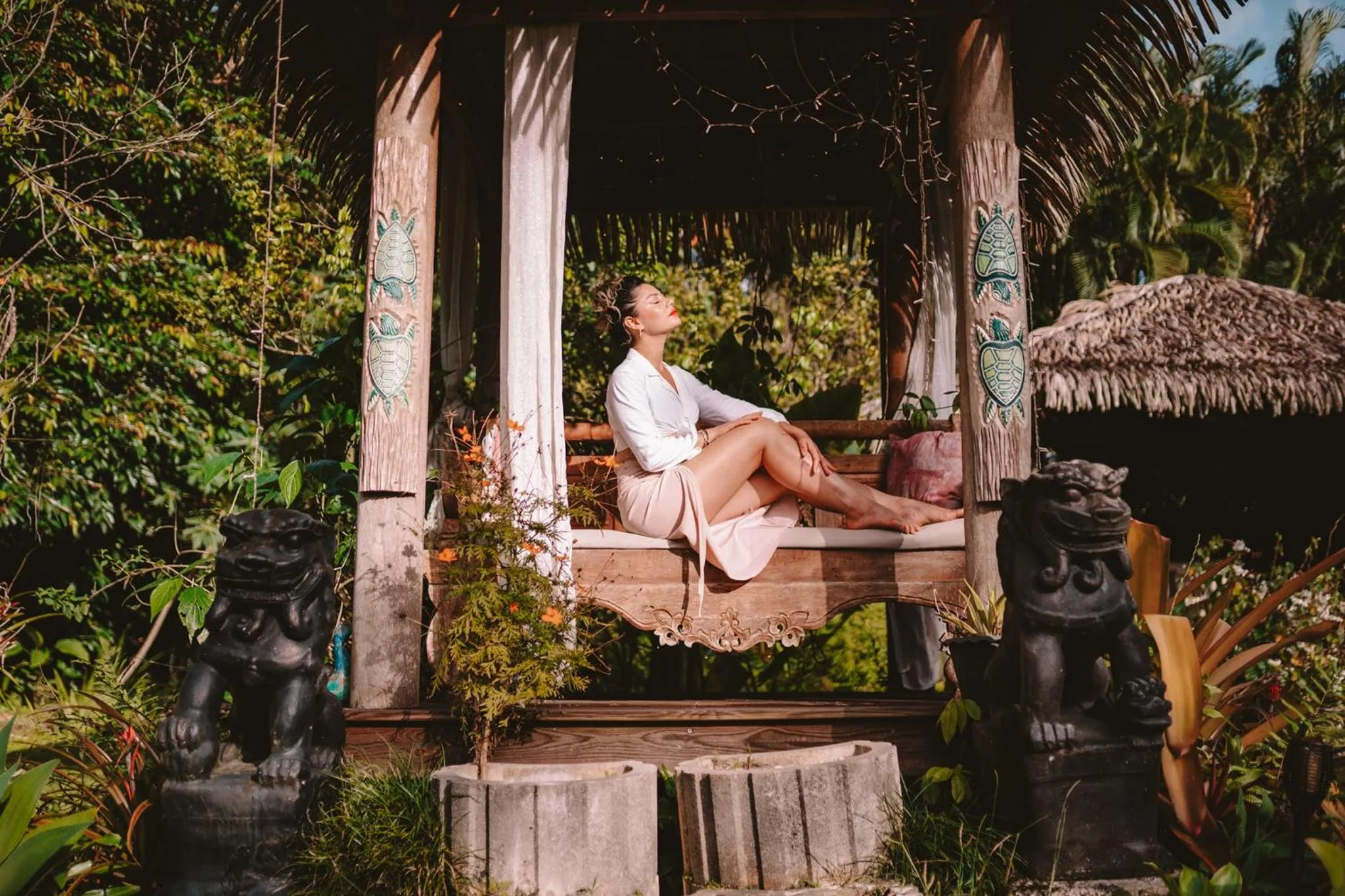 Une femme se repose sur une banquette en bois sculpté dans un jardin tropical.