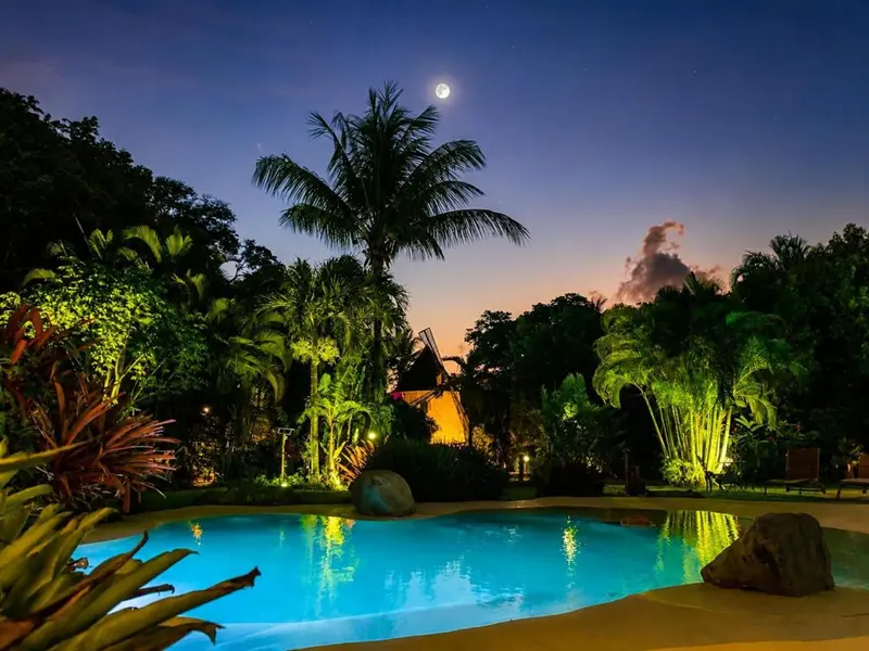 Piscine illuminée au crépuscule, végétation tropicale en arrière-plan et pleine lune.