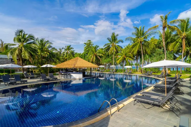Une piscine bleue bordée de chaises longues et de parasols, avec une végétation tropicale en arrière-plan.