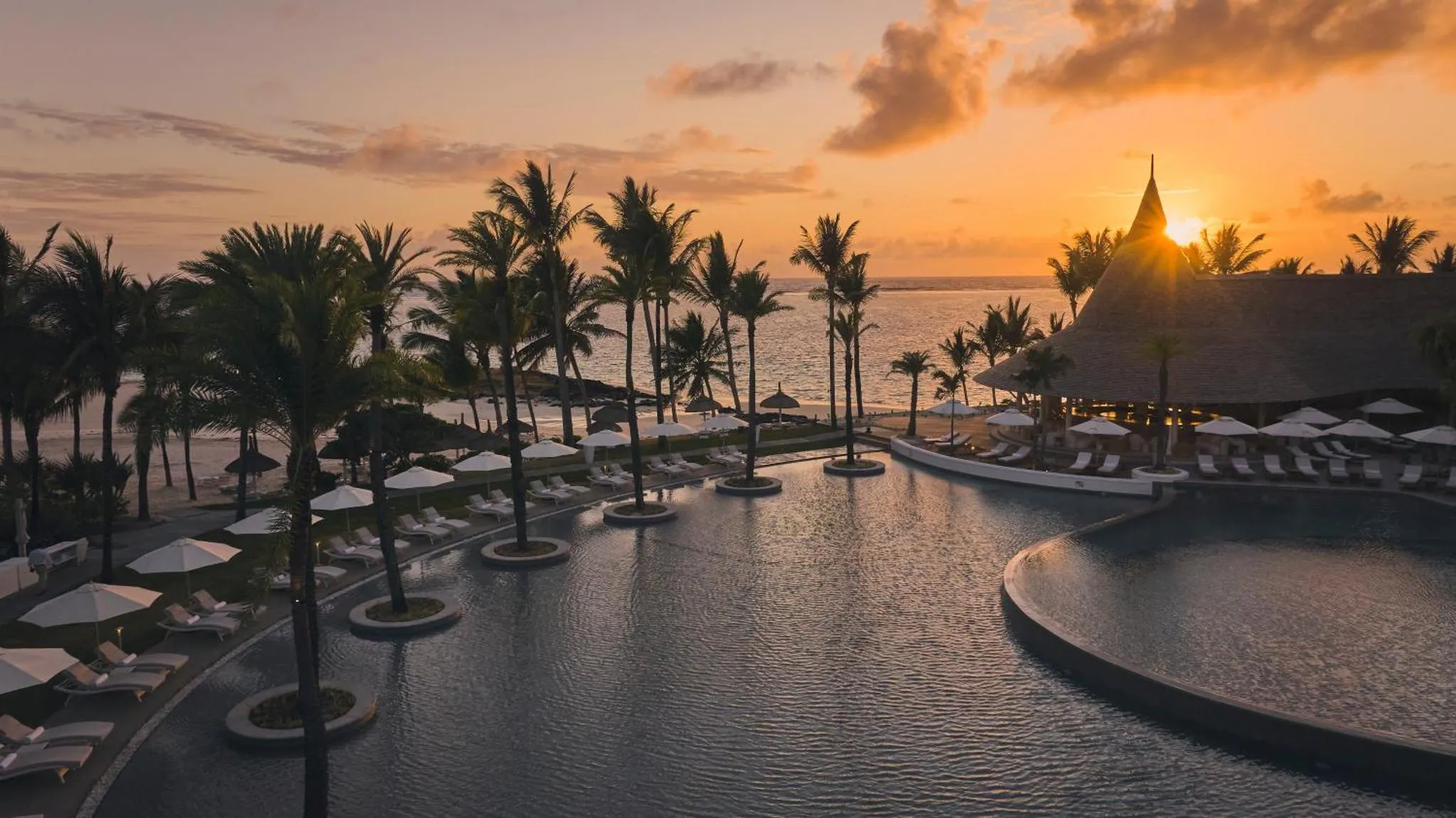Une piscine bordée de transats et de palmiers au coucher du soleil.