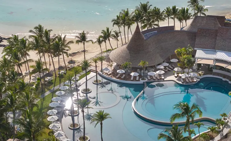 Vue aérienne d'une piscine sinueuse bordée de palmiers et d'un bâtiment à toit de chaume près d'une plage.