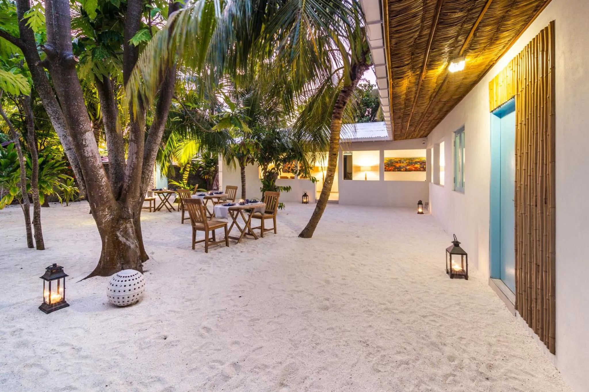 Terrasse ombragée avec tables et chaises en bois sur sable blanc.