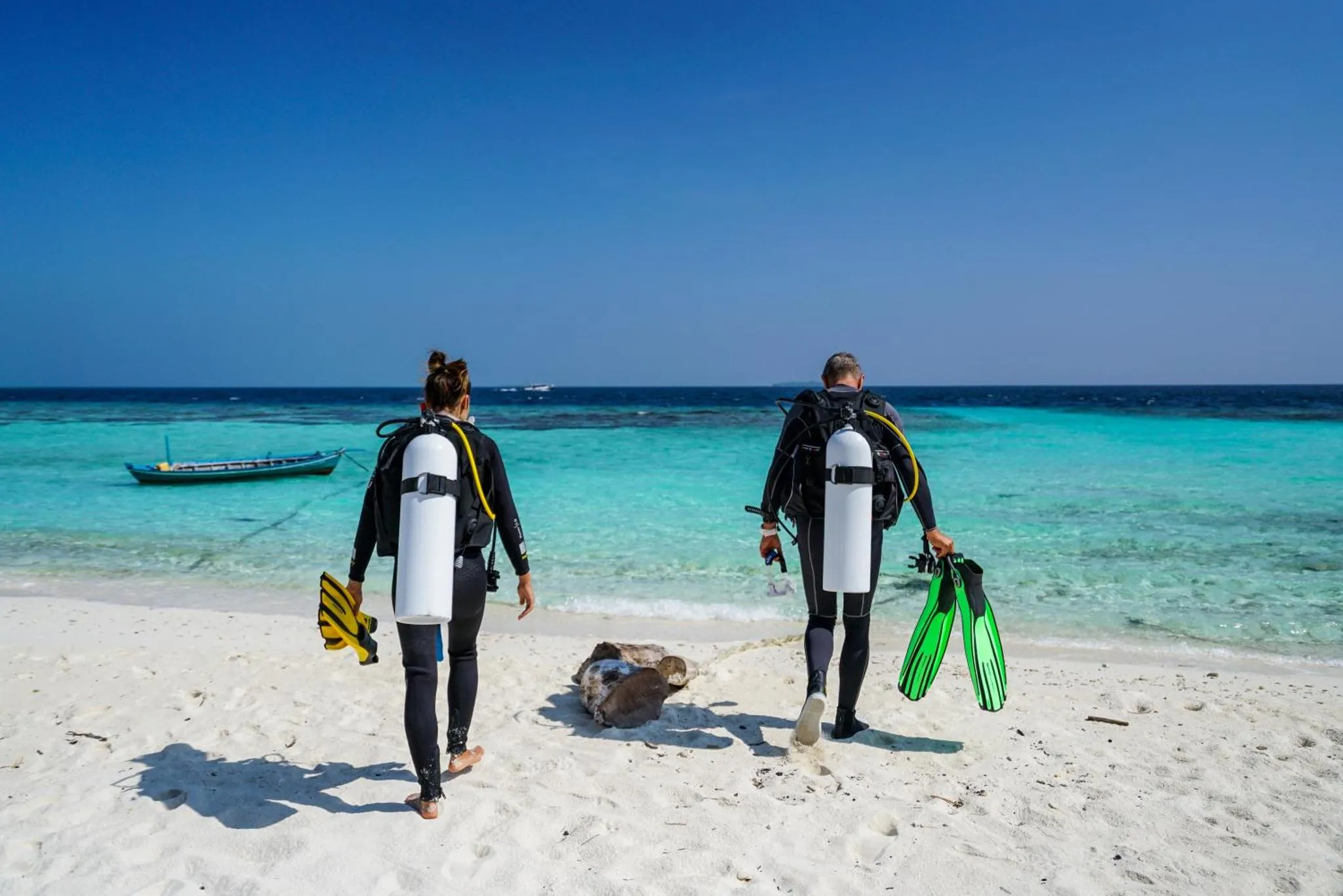 Deux personnes en combinaison de plongée marchent sur une plage de sable blanc vers une mer turquoise.