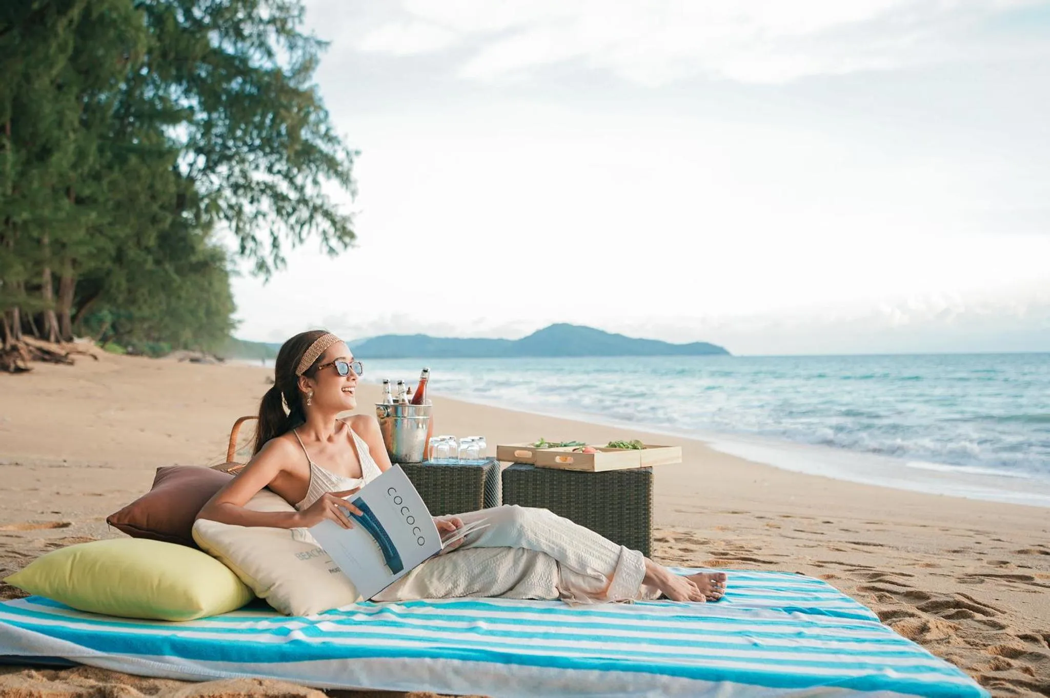 Une femme se détend sur une plage de sable fin avec une boisson et un livre.