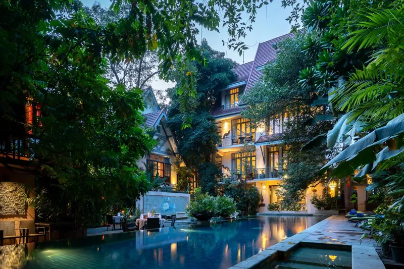 Une piscine illuminée au milieu d'une végétation luxuriante, avec un bâtiment éclairé en arrière-plan.