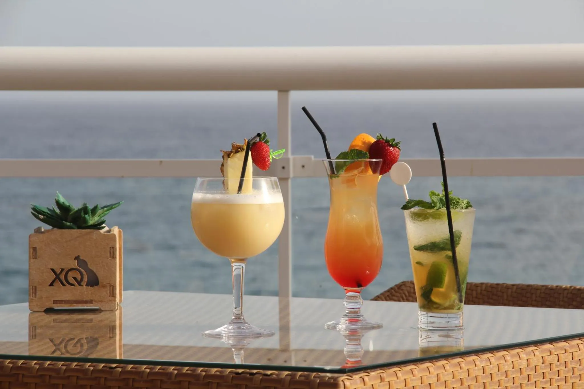 Trois cocktails colorés sont présentés sur une table en osier avec vue sur la mer.