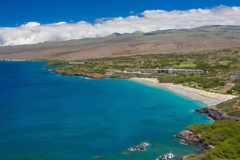 Vue aérienne d'une plage de sable fin bordée d'une eau turquoise et d'une végétation luxuriante, avec des bâtiments en arrière-plan.