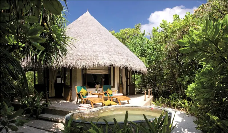 Une terrasse en bois avec des fauteuils et un petit bassin d'eau devant une villa tropicale.