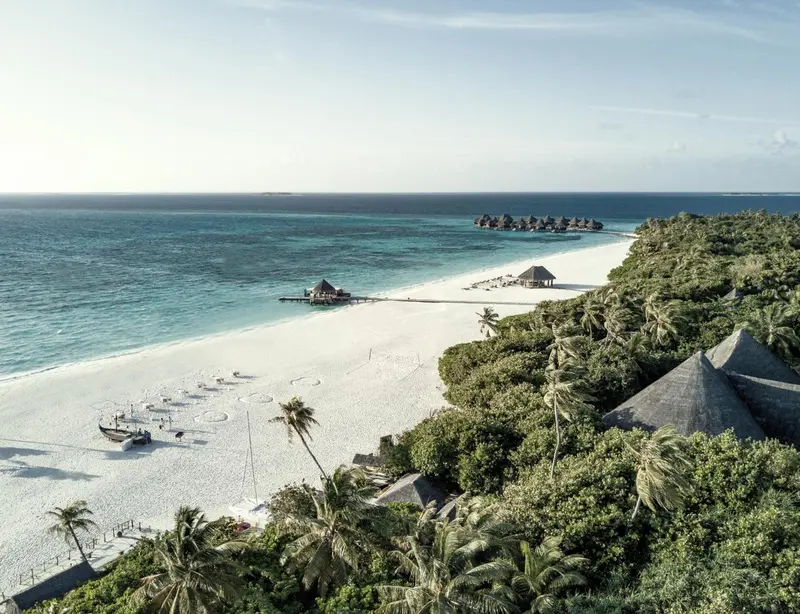 Vue aérienne d'une plage de sable blanc bordée de végétation luxuriante et de bungalows sur pilotis.
