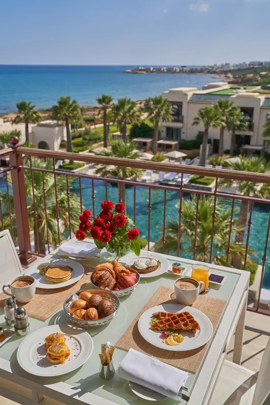Une table de petit-déjeuner est dressée sur une terrasse avec vue sur la mer et la végétation.