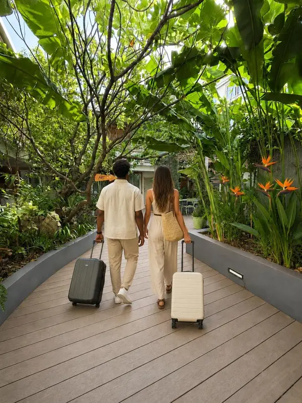 Un couple marchant sur une allée bordée de végétation tropicale, chacun tirant une valise.