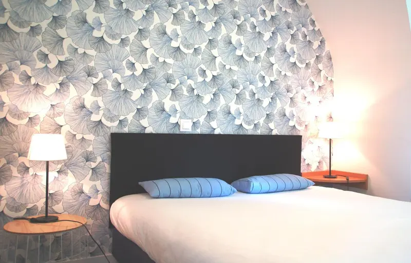 Une chambre avec un lit recouvert d'une courtepointe blanche et de coussins bleus, face à un mur décoré d'un motif floral.