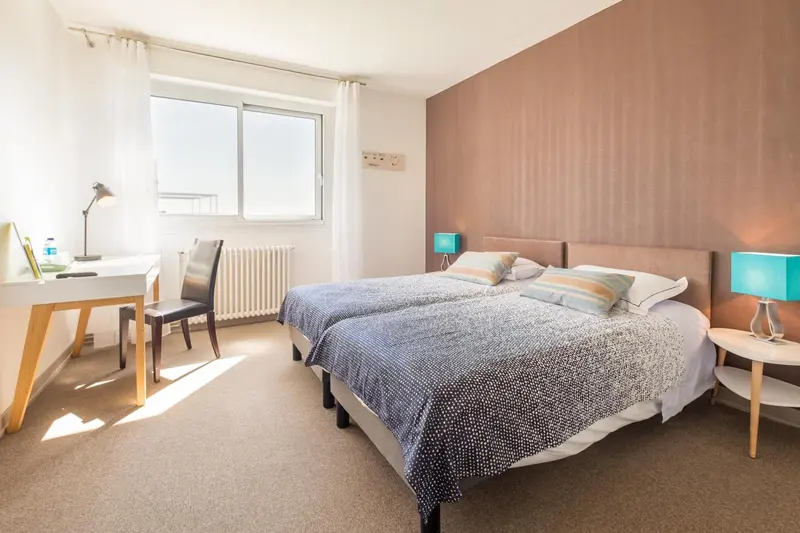 Une chambre lumineuse avec deux lits simples et un bureau près de la fenêtre.