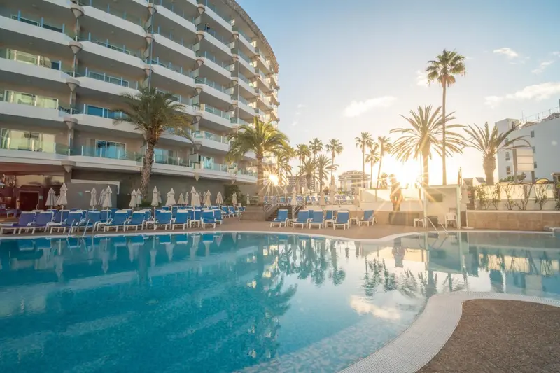 Vue d'une piscine devant un bâtiment moderne bordé de palmiers au coucher du soleil.