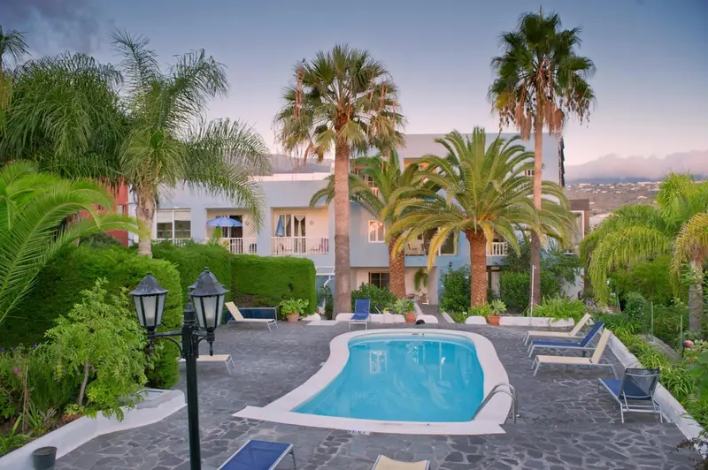 Une piscine turquoise est entourée de chaises longues et de végétation luxuriante devant un bâtiment clair.