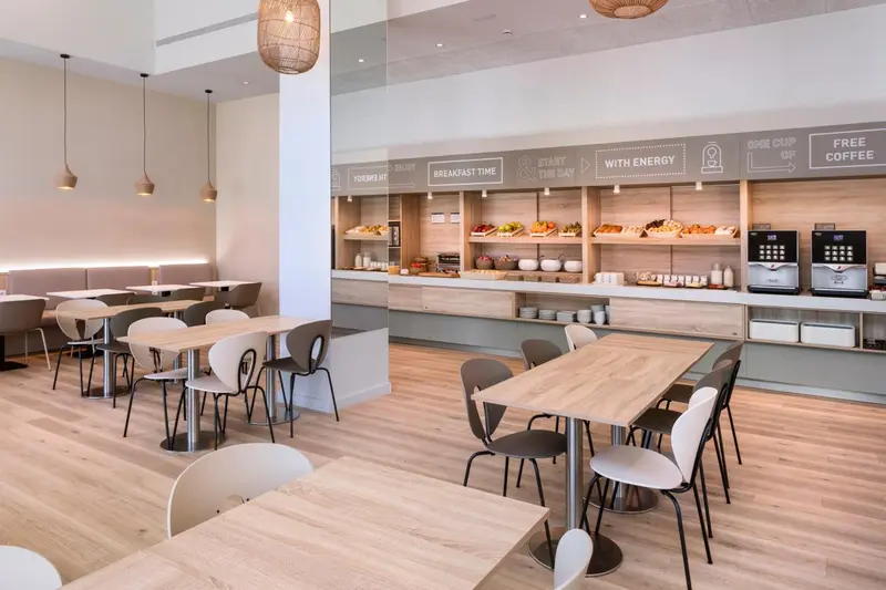Une salle lumineuse avec des tables et des chaises, un buffet de petit-déjeuner et des suspensions décoratives.
