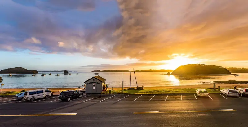 Un parking en bord de mer au coucher du soleil, avec des bateaux amarrés au loin.