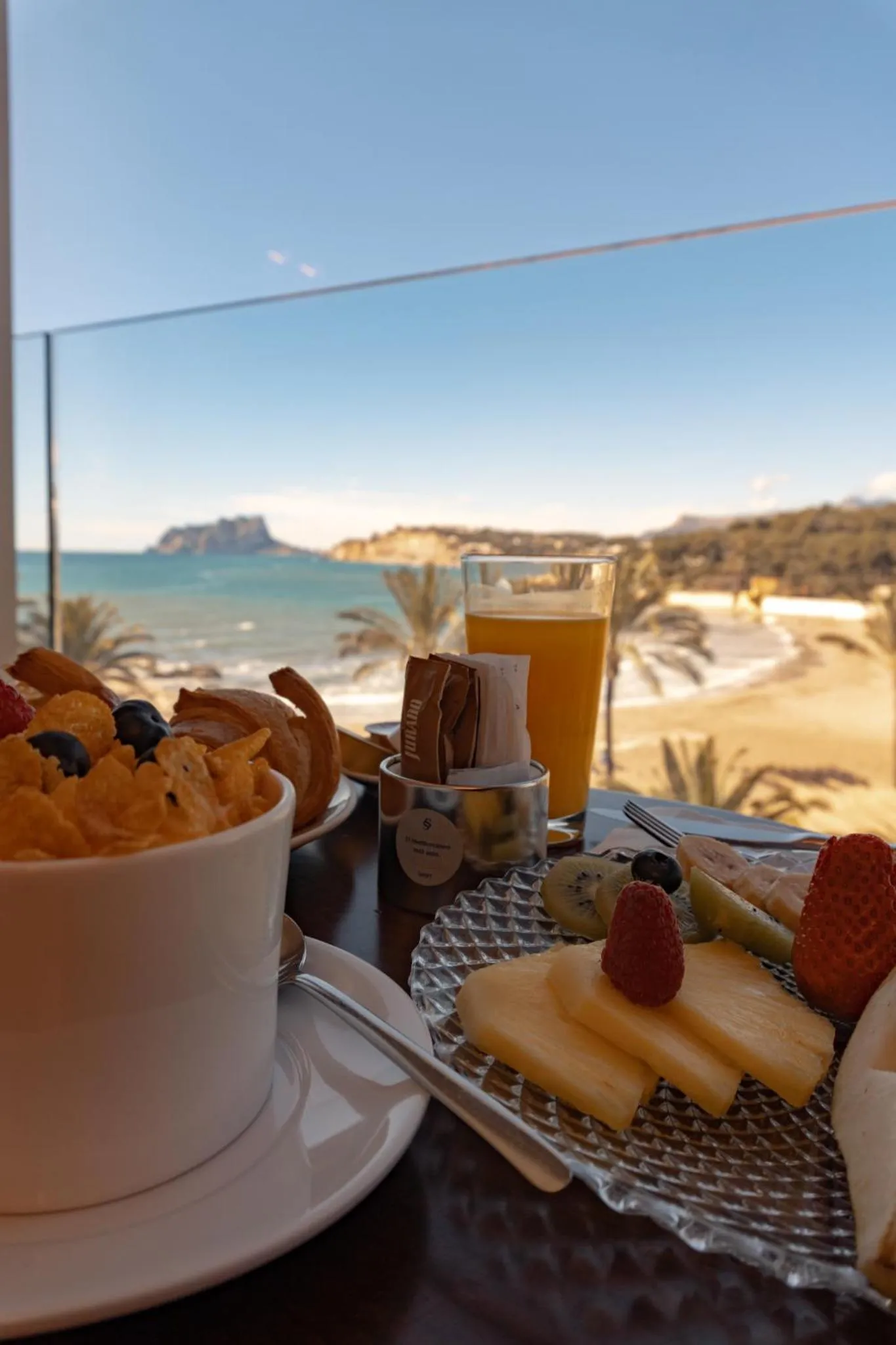 Un petit-déjeuner avec des fruits et une boisson est présenté sur une table avec vue sur la mer et une plage.