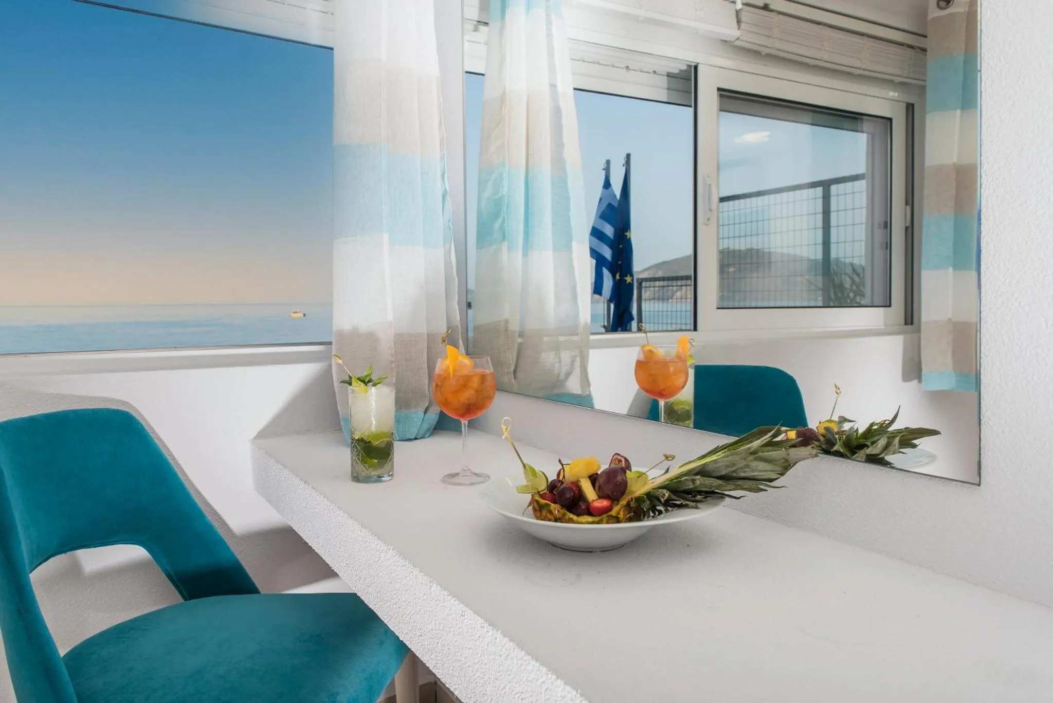 Une table est dressée avec des boissons et des fruits devant une grande fenêtre donnant sur la mer.