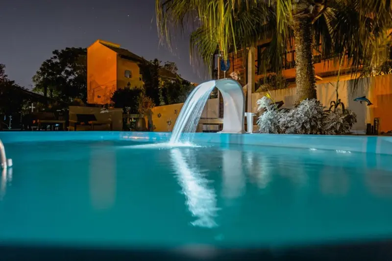 Une piscine illuminée avec une cascade artificielle et des palmiers.
