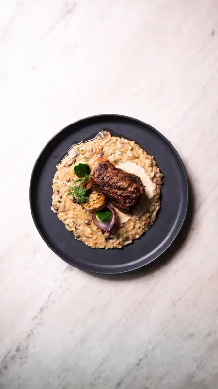 Assiette de risotto crémeux surmontée d'une pièce de viande grillée et de quelques légumes.