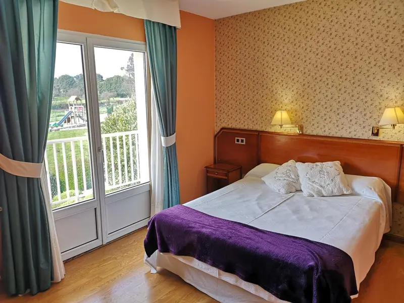 Chambre lumineuse avec un lit recouvert d'une courtepointe violette et une vue sur un espace extérieur.