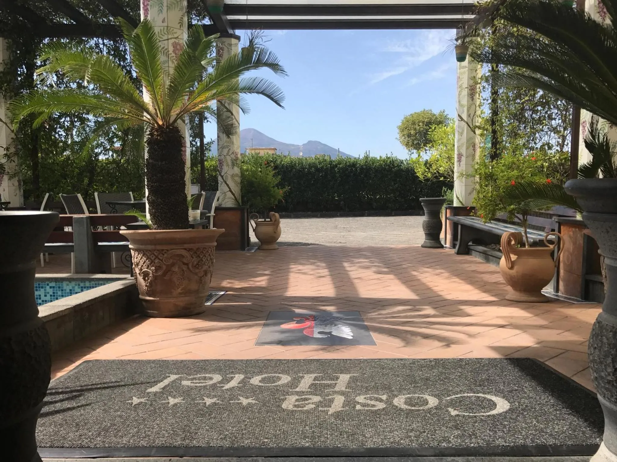 Une entrée d'établissement s'ouvre sur une cour ombragée avec des plantes et une piscine.