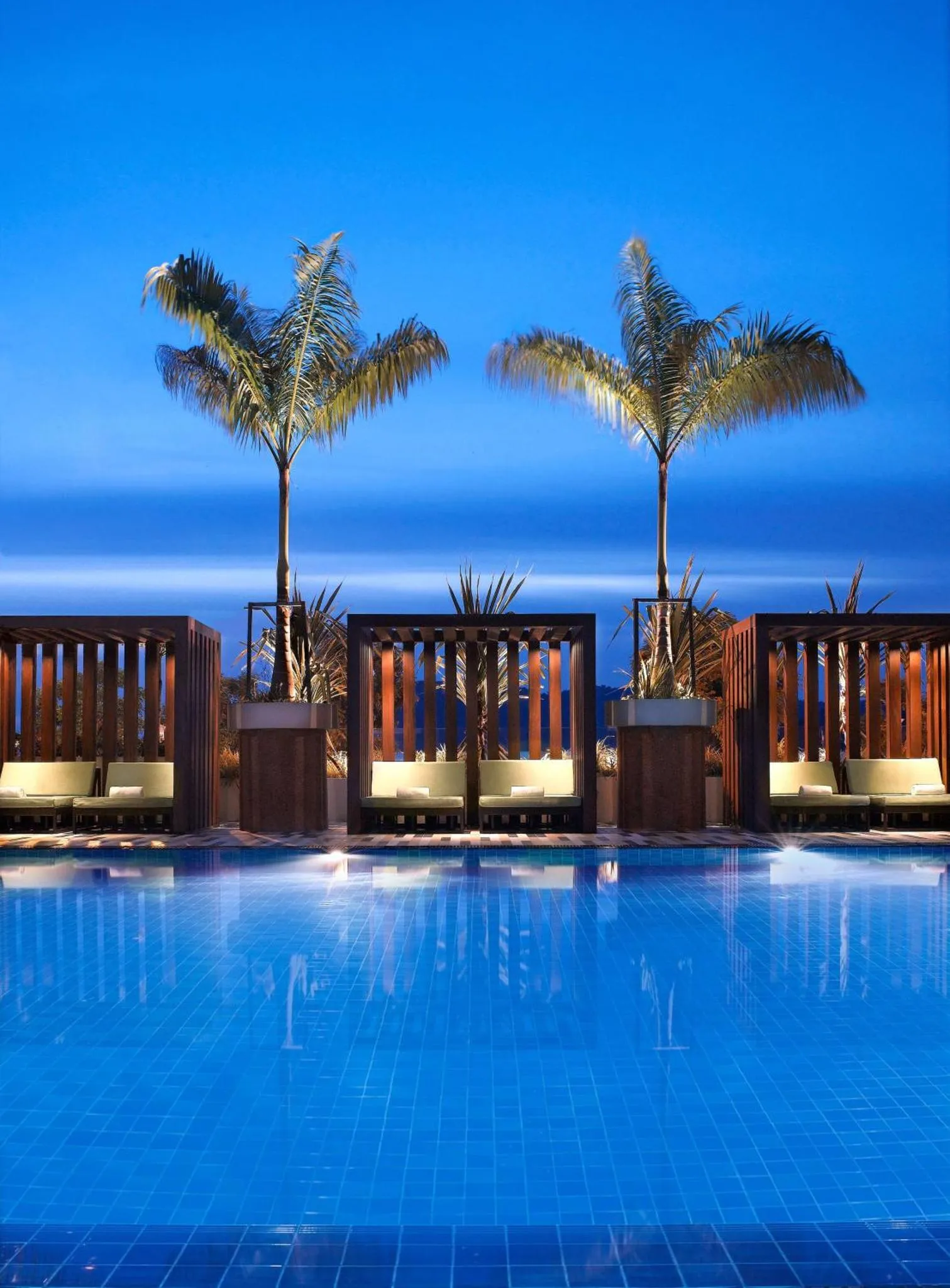 Trois lits de repos bordent une piscine illuminée sous un ciel bleu foncé.