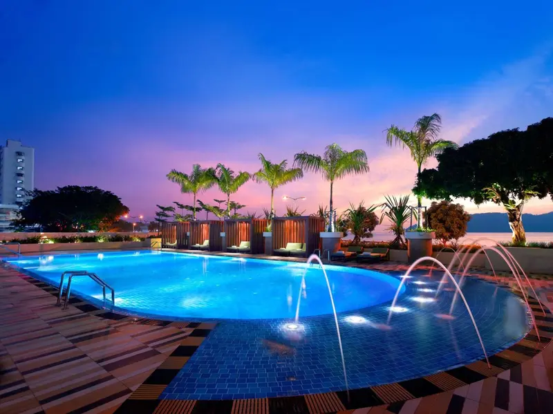 Piscine illuminée avec jets d'eau et végétation tropicale au coucher du soleil.