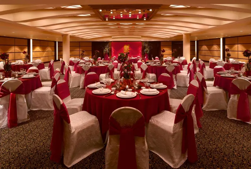 Une grande salle de réception est mise en scène avec des tables rondes nappées de rouge et de blanc pour un événement festif.