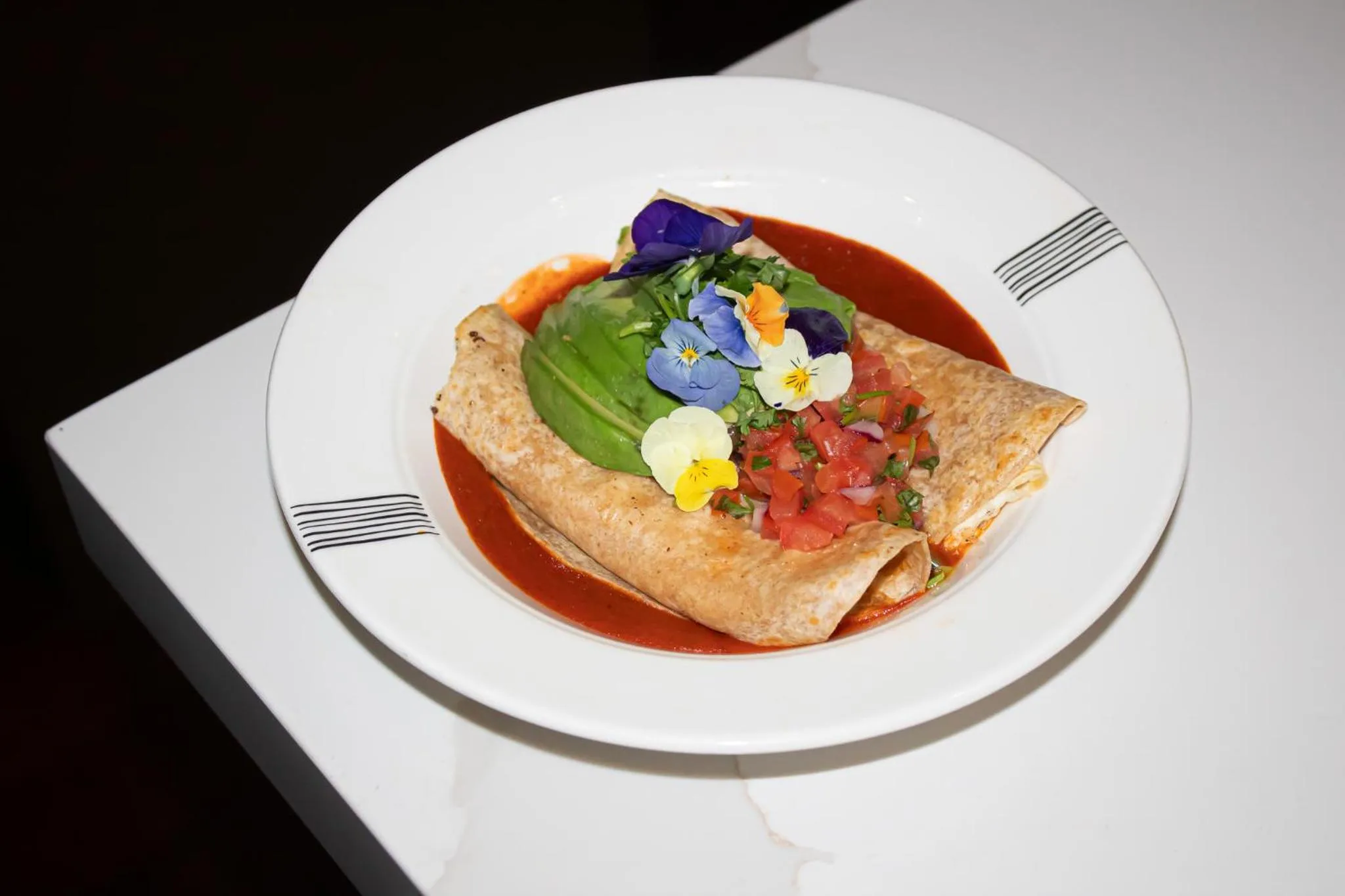 Assiette présentée avec un plat enroulé, garni de sauce, d'avocat, de tomates et de fleurs comestibles.