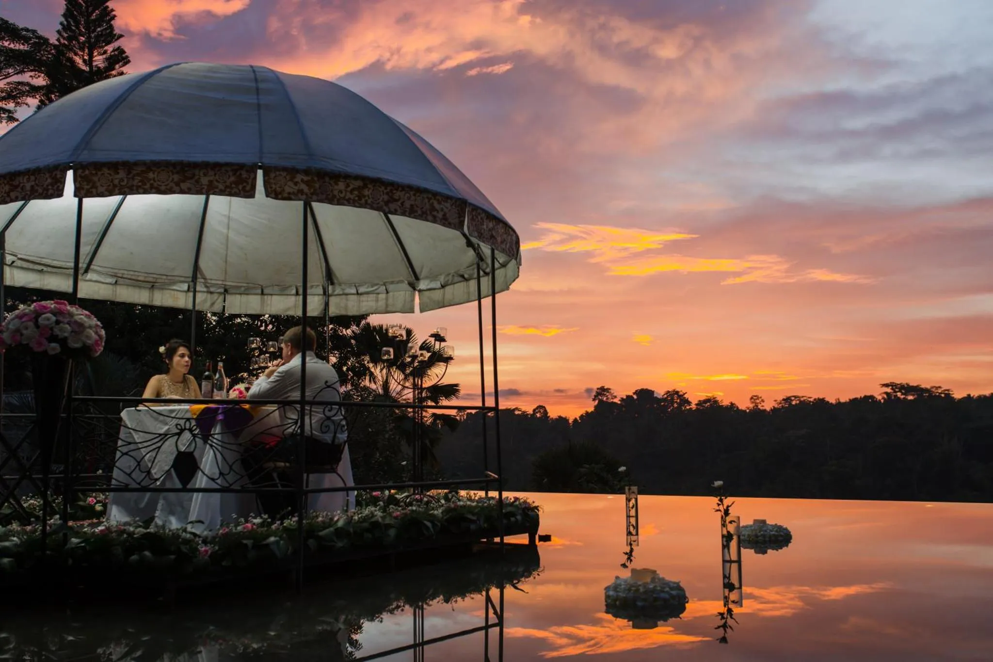 Un couple se tient dans un gazebo au bord d'une piscine reflétant un ciel orangé.