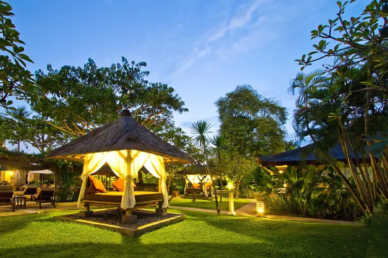 Un pavillon illuminé se dresse sur une pelouse verdoyante, entouré de végétation tropicale.
