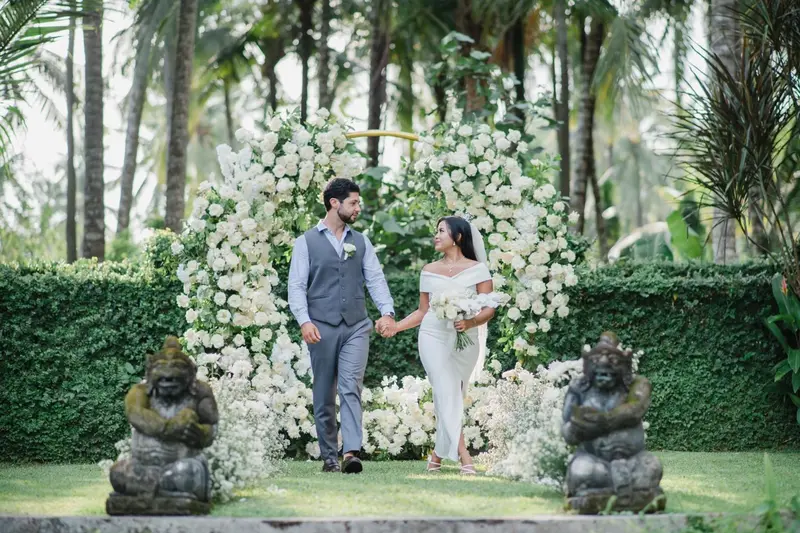 Un couple se promène main dans la main dans un jardin luxuriant orné de fleurs blanches et de statues.