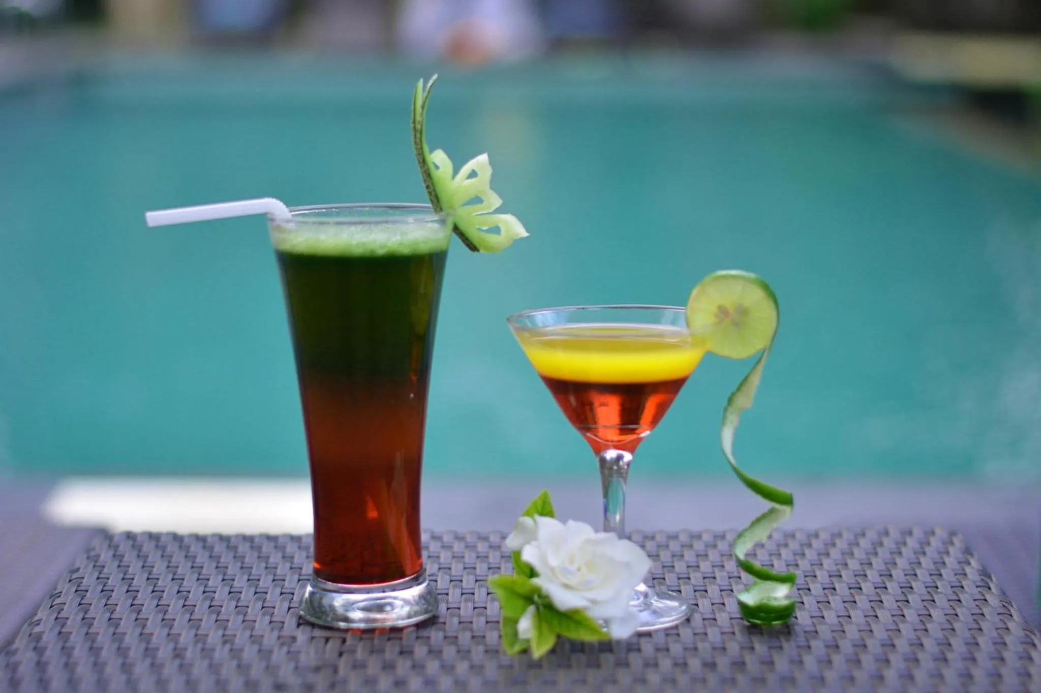 Deux boissons colorées sont présentées sur une table en osier devant une piscine.