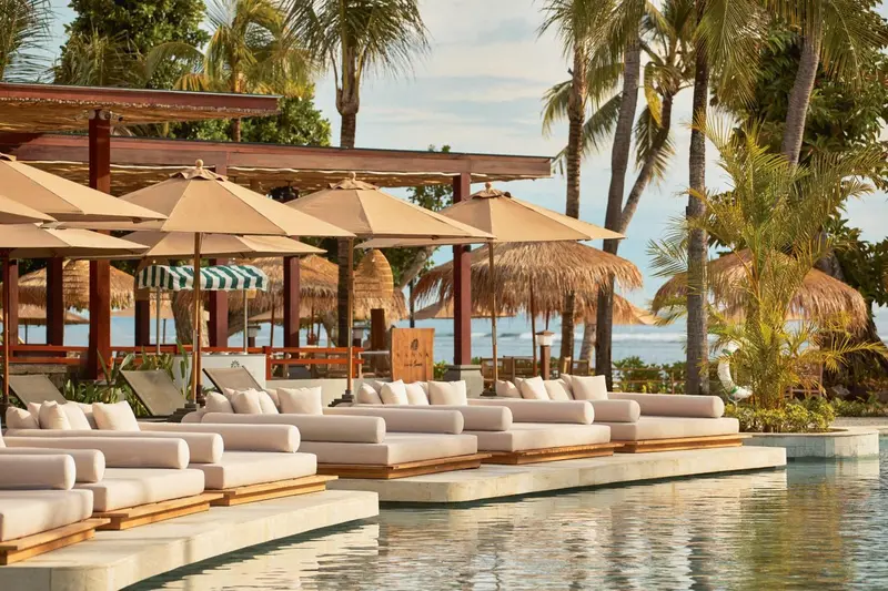Transats et parasols beige disposés au bord d'une piscine turquoise, avec des palmiers en arrière-plan.