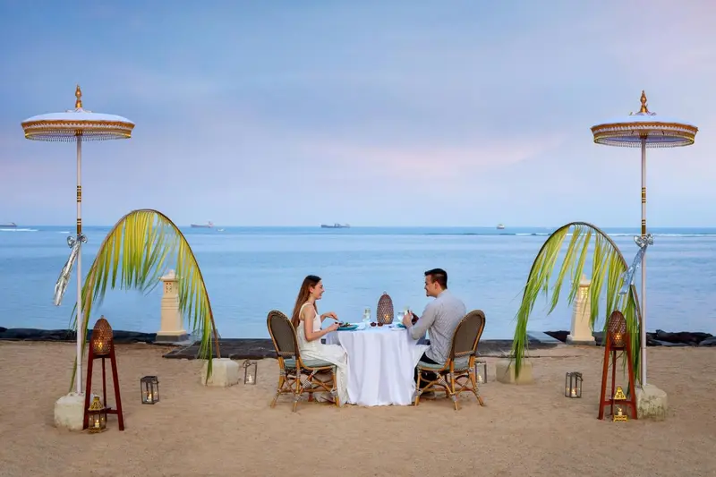 Un couple dîner à une table dressée sur une plage de sable au coucher du soleil.