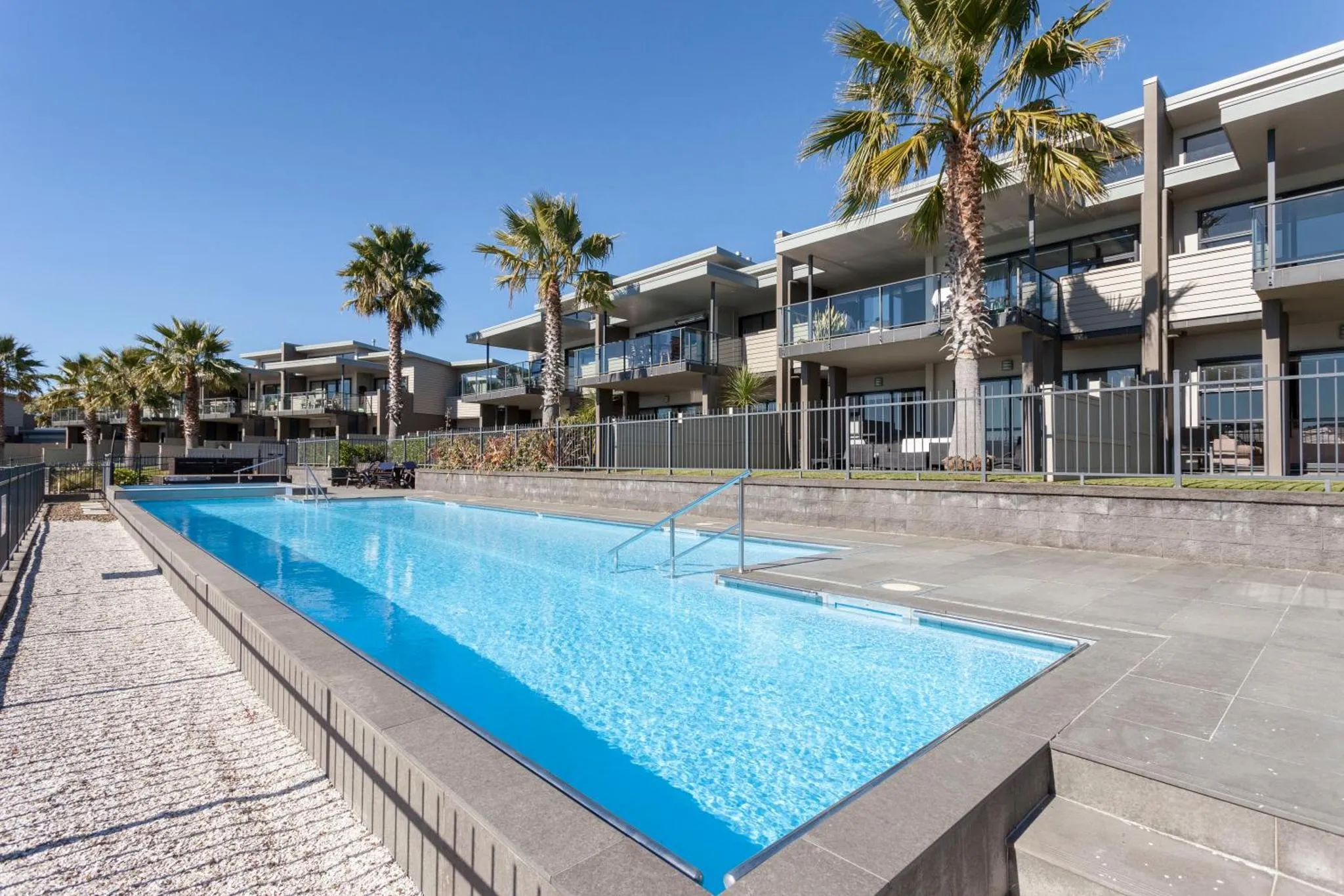 Une piscine rectangulaire bordée d'une terrasse ensoleillée devant un bâtiment moderne.