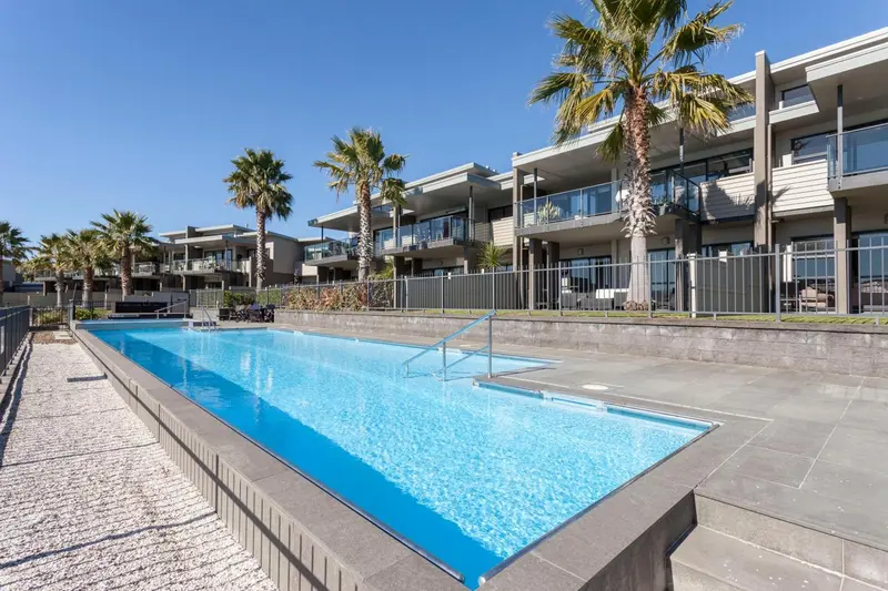 Une piscine rectangulaire bordée d'une terrasse ensoleillée devant un bâtiment moderne.