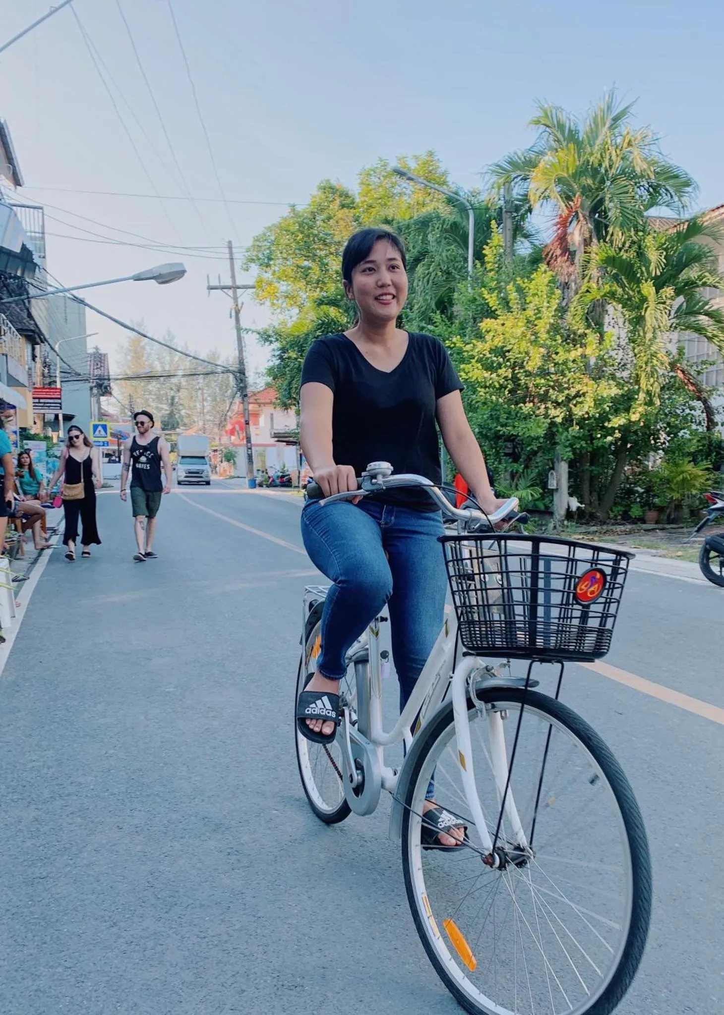 Une femme souriante fait du vélo sur une route bordée d'arbres et de bâtiments.