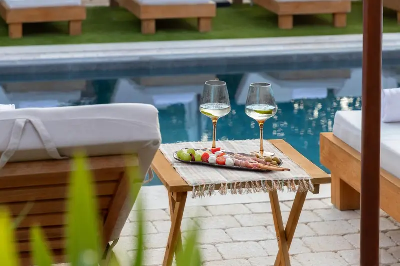 Deux verres de vin et une assiette de charcuterie sont posés sur une table basse au bord d'une piscine.
