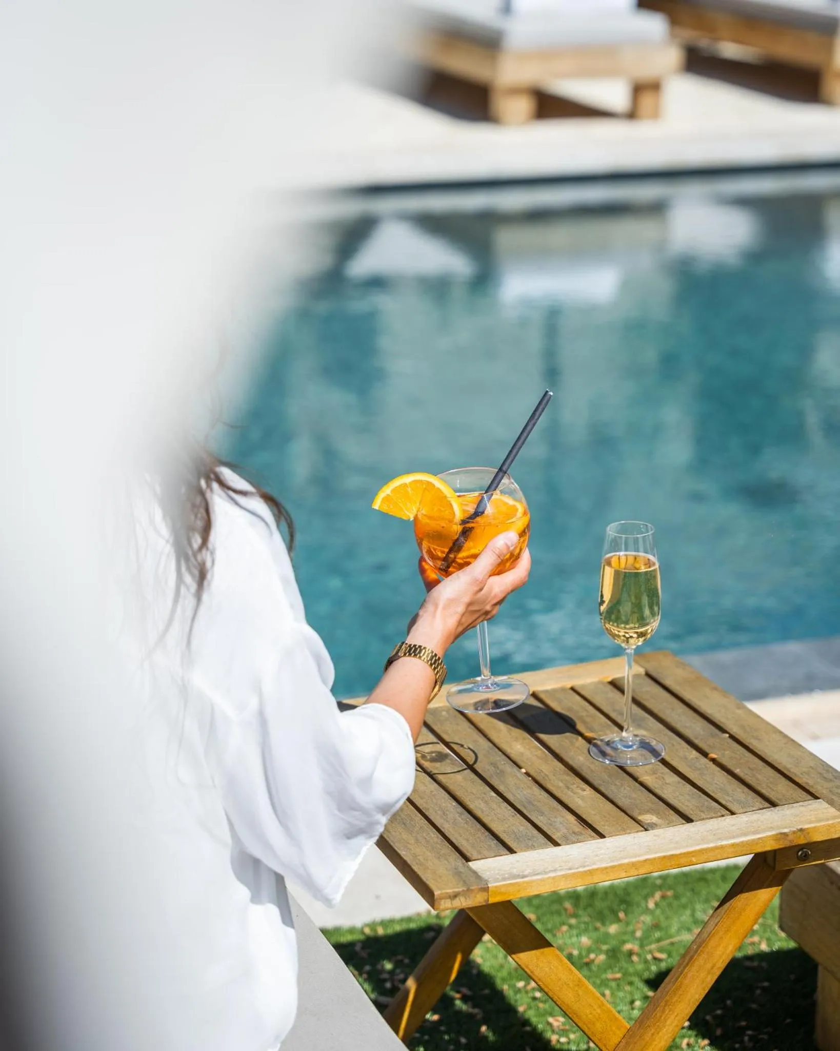 Une personne tient un cocktail devant une piscine ensoleillée.