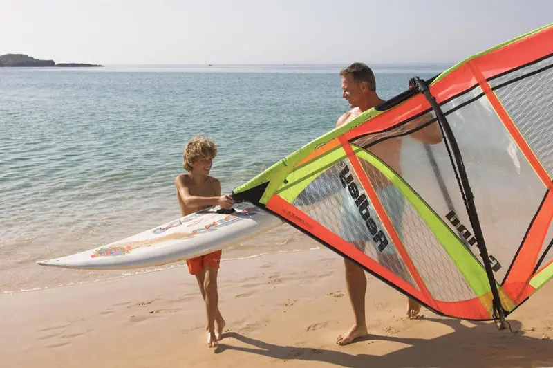 Un homme et un enfant portent du matériel de planche à voile sur une plage.